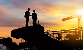 Silhouette of two construction professionals on a jobsite at sunset with cranes in the background, illustrating ABC’s August 2025 report showing backlog at 8.5 months and contractor optimism holding steady.