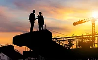 Silhouette of two construction professionals on a jobsite at sunset with cranes in the background, illustrating ABC’s August 2025 report showing backlog at 8.5 months and contractor optimism holding steady.