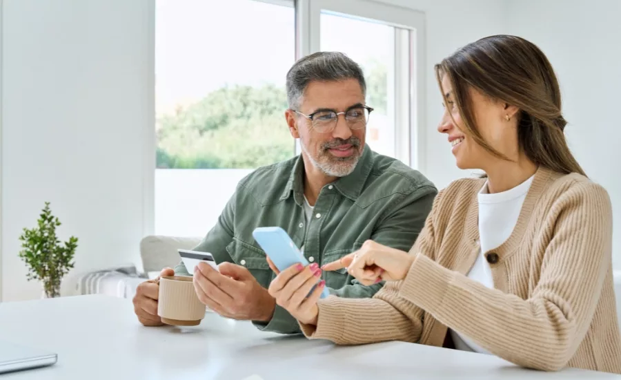 Happy mature couple using phone holding credit card making payments. Middle aged older man and woman doing ecommerce shopping on smartphone booking or buying online on mobile sitting at home table.