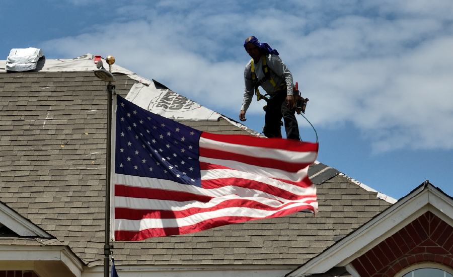 A roofing contractor stands on a roof behind an American flag