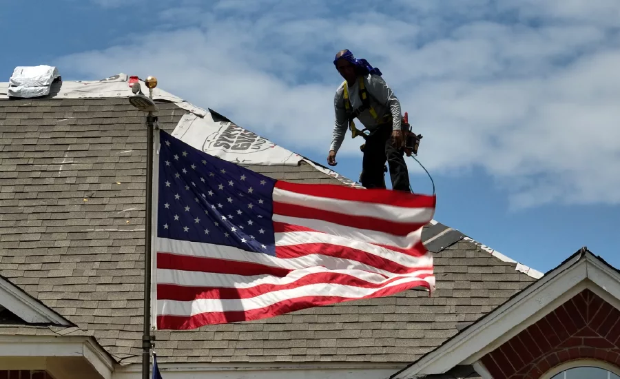 A roofing contractor stands on a roof behind an American flag