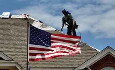 A roofing contractor stands on a roof behind an American flag