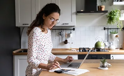 Female standing in home kitchen doing accounting work from home calculates company financial statement, use calculator fill data results on laptop application, housewife manage family budget