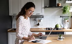 Female standing in home kitchen doing accounting work from home calculates company financial statement, use calculator fill data results on laptop application, housewife manage family budget