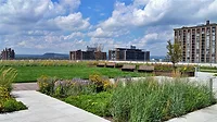 Rooftop garden with green lawn, flowerbeds, benches, and distant buildings under a cloudy sky.